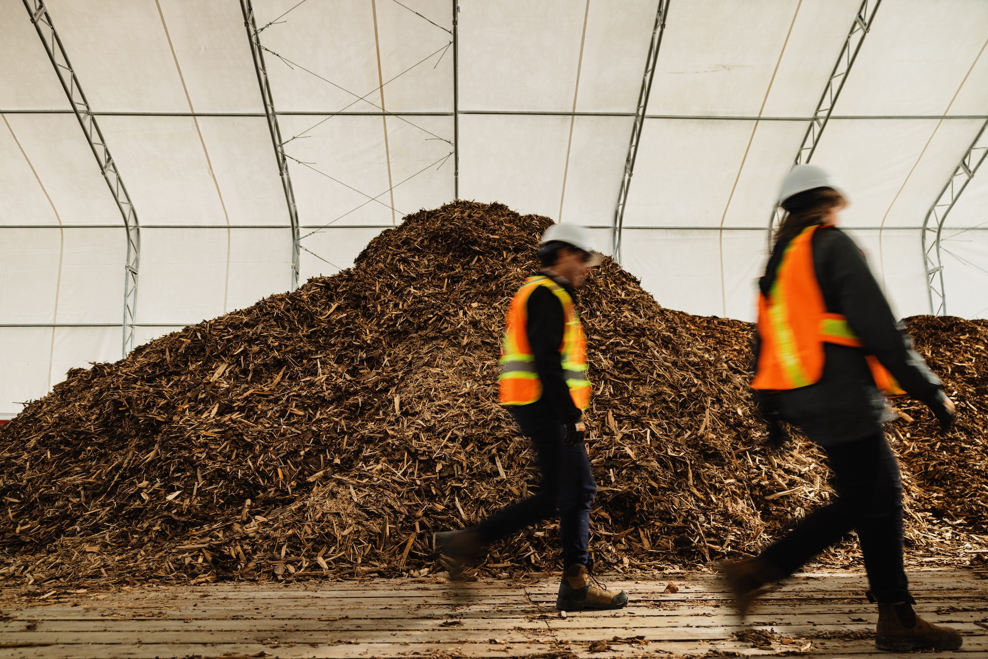 Two people walk past a feedstock wood chip pile