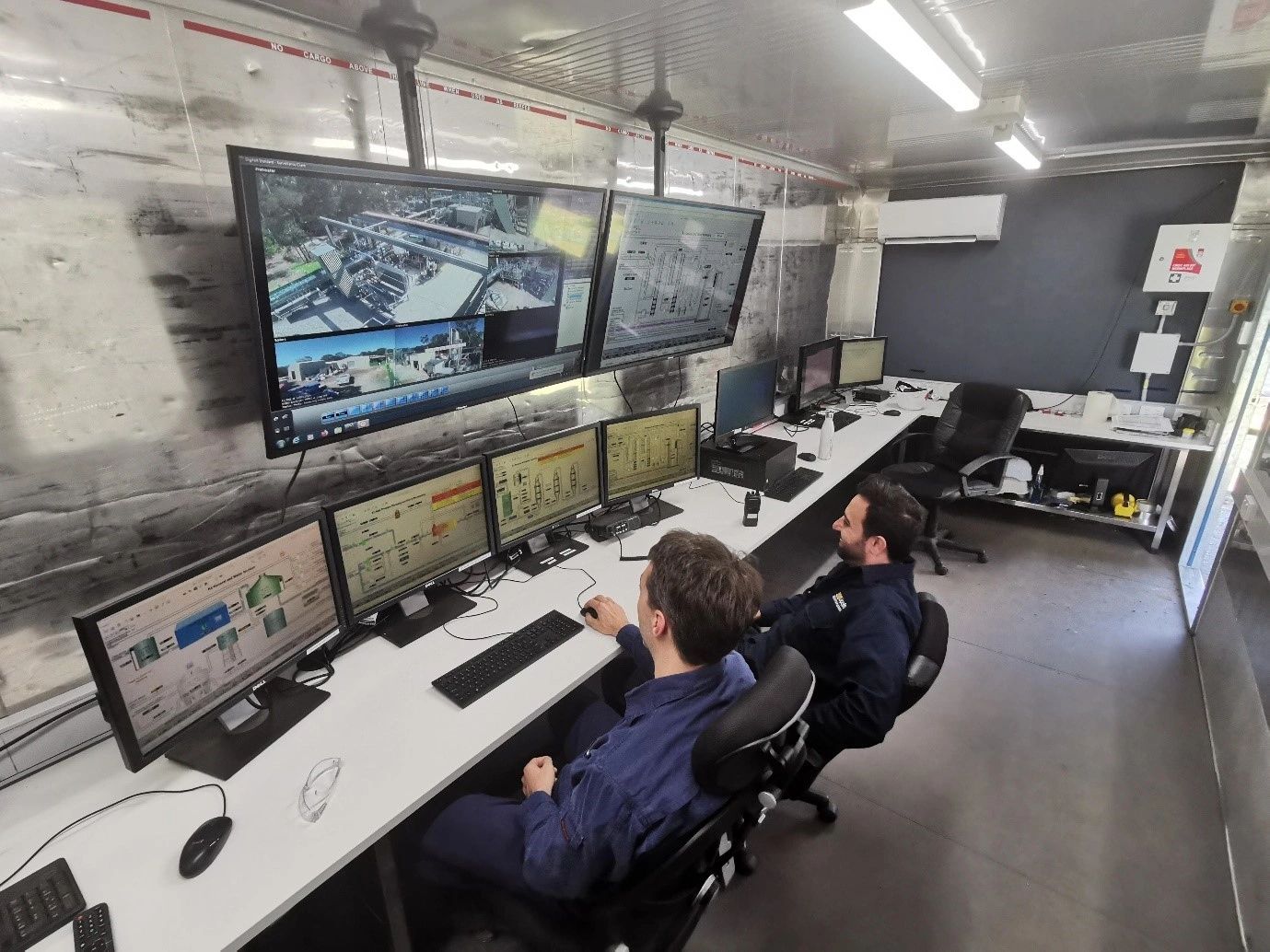 Two men sit at a desk in front of many monitors